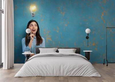 Illuminated light bulb above the head of a young woman, the concept of having an idea of a surprised girl with eureka on an old blue wall  Wall mural