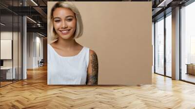 Smiling blond pretty smiling girl beauty female gen z model with short blonde hair beautiful face healthy skin and tattoo looking at camera wearing white top isolated at beige background Wall mural