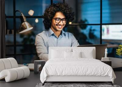 Smiling young curly indian latin ethnic business man or student wearing glasses remote working overtime, learning online late at night at home or in dark office using laptop computer at workplace Wall mural