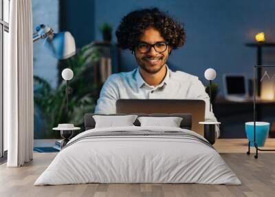 Smiling young curly indian latin ethnic business man or student wearing glasses remote working overtime, learning online late at night at home or in dark office using laptop computer at workplace Wall mural