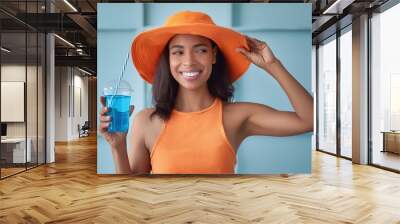 Stylish young woman in a vibrant orange hat and tank top holding a cool blue beverage Wall mural