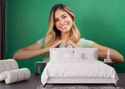 Young happy latin woman showing heart making shape with hands isolated on green background. Smiling female model expressing love and dating romance, warm affection, health care concept Wall mural