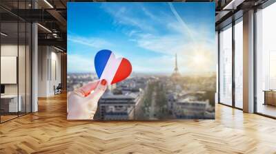 Girl's hand holding heart with french flag colors. Heart is on background of historic city center of Paris, Eiffel Tower, blue sky, clouds, sun. Love, Paris, travel concept. Wall mural