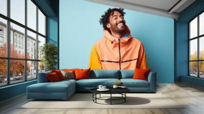 Portrait of a joyful indian man in his 20s wearing a functional windbreaker in pastel orange background Wall mural