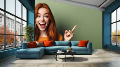 An excited young ginger teenage girl smiling and pointing her index finger up at the copy space expressing wow emotion standing isolated on green background studio copy space, advertisement concept Wall mural