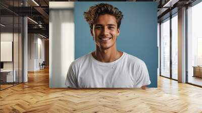 Young man wearing a white t-shirt against a blue background looks confident with a smile on his face Wall mural