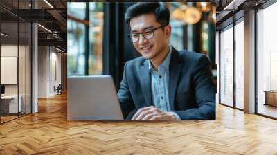 Asian businessman typing on laptop during work in cafe. Concept of remote and freelance work. Smiling adult successful man wearing suit and glasses sitting at wooden desk. Sunny day Wall mural