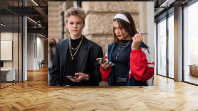 Cute fun curly teenage boy and girl wearing stylish 90s fashion clothes with chains around neck. Youth subculture. Happy teenage boy and girl wears stylish clothes with chains with phone for a walk. Wall mural