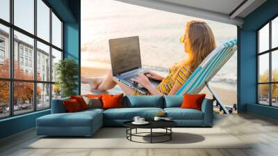 Fit young woman sitting on deck chair at sea view beach using laptop. Female freelance programmer in chaise-long lounge working coding surfing on notebook computer, blank screen. Remote work concept. Wall mural