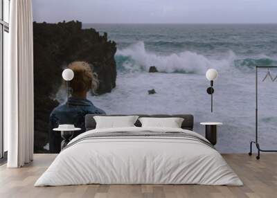 Young woman standing next to the ocean and looking at rough sea and breaking waves Wall mural