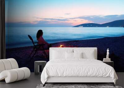 Silhouette of a young woman sitting in the evening by the fire on the shore of the Mediterranean Sea.  Wild camping, freedom, van life. Travelling in a camper van. Independence and comfort. Wall mural