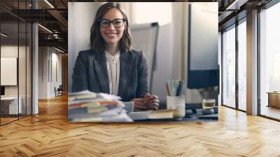 Headshot of attractive business woman in glasses, smiling while sitting in front of her computer, looking into the camera. Wall mural