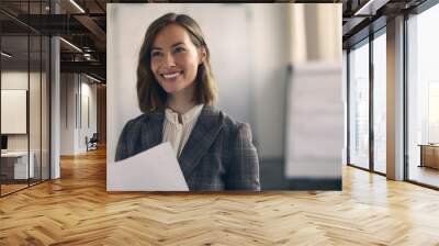 Portrait of smiling pretty business woman standing in the office with some paperwork	 in hands looking at the colleague she is talking to. Happy coworker and satisfied about the job.  Wall mural