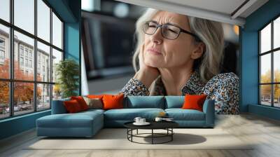 A middle aged woman is massaging her stiff neck while sitting at her desk, showing signs of discomfort and stress. This image captures struggle of managing work related tension and need for self care Wall mural