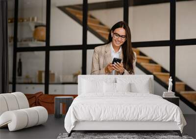 woman sitting on sofa in office relaxed using smartphone in beige jacket and jeans listening to earpods Wall mural