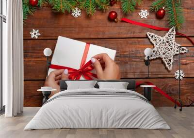 Female hands in a red new year sweater holding a white gift box on a brown wooden background, fir tree, christmas baubles, toys. Flat lay. Festive background. Wall mural