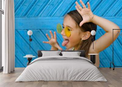 A brunette girl in yellow sunglasses stands against a blue wall of planks and makes a funny grimace, showing her tongue. Naughty ill-mannered child Wall mural