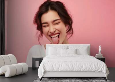 Smiling girl winks at the camera with tongue out in a playful pose. Pink background highlights her happiness and cheerful expression. Young adult caucasian woman shows a casual carefree attitude. Wall mural