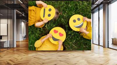 Three children lying on the grass are holding cardboard emoticons with different emotions in their hands: a sad, smiling happy smile, a loving smiley with hearts instead of eyes.  World Emoji Day Wall mural