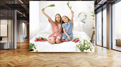 Two girls happily waving tulips sitting in bed. world children's day. Wall mural