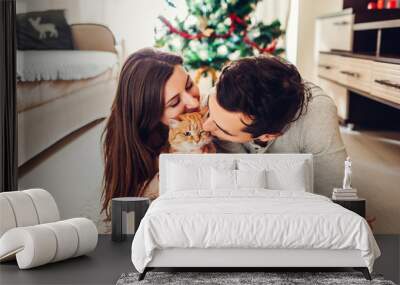 Couple in love lying by Christmas tree and playing with cat at home. Man and woman relaxing Wall mural