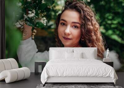 Portrait of young attractive woman posing in white shirt walking in blooming garden among flowers. Girl enjoys nature Wall mural