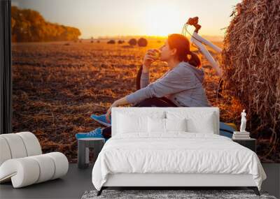 Young bicyclist having rest after a ride in autumn field at sunset. Woman eating by haystack Wall mural