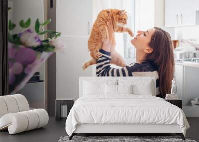 Young woman playing with cat in kitchen at home. Girl holding and red raising cat Wall mural