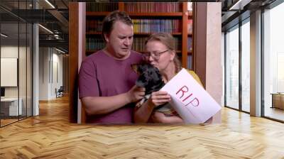 Young married couple leaves room with dachshund and piece of paper with inscription RIP in their hands. Owners found out bad news about health of their beloved dog, they are upset and in tears. Wall mural