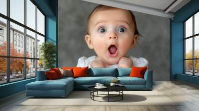 Surprised baby with wide eyes and open mouth, expressing shock or awe against a gray background. Wall mural