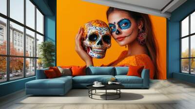 Halloween or Day of the Dead. Cheerful woman with mexican style face art posing with decorative skull on orange background in studio. Woman with bright Wall mural