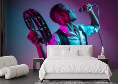 African American handsome jazz musician playing tambourine and singing into the microphone in the studio on a neon background. Music concept. Young joyful attractive guy improvising. Close-up retro Wall mural