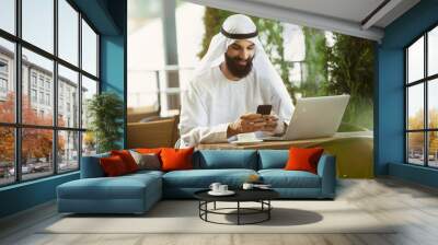 Arab saudi businessman working online with a laptop and tablet in a coffee shop or a cafe with an outdoor terrace in the background. Concept of business, finance, modern technologies, start up. Wall mural