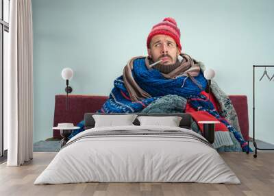 Bearded sick man with flue sitting on sofa at home or studio with thermometer covered with knitted warm clothes. Illness, influenza concept. Relaxation at Home. Healthcare Concepts. Wall mural