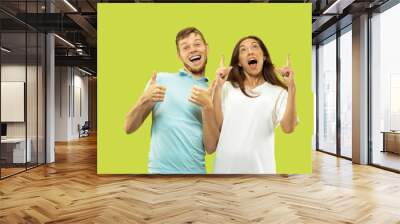 Beautiful young couple's half-length portrait isolated on green studio background. Woman and man standing and pointing up, showing the sing of OK. Facial expression, human emotions concept. Wall mural