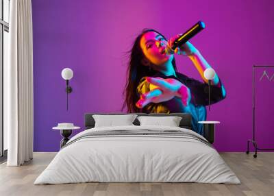 Caucasian female singer portrait isolated on purple studio background in neon light. Beautiful female model in black wear with microphone. Concept of human emotions, facial expression, ad, music, art. Wall mural