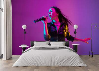 Caucasian female singer portrait isolated on purple studio background in neon light. Beautiful female model in black wear with microphone. Concept of human emotions, facial expression, ad, music, art. Wall mural