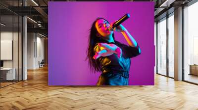 Caucasian female singer portrait isolated on purple studio background in neon light. Beautiful female model in black wear with microphone. Concept of human emotions, facial expression, ad, music, art. Wall mural