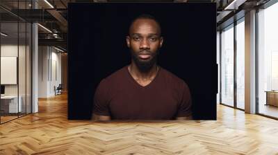 Close up portrait of young african man isolated on black studio background. Photoshot of real emotions of male model. Standing and looks serious. Facial expression, human nature and emotions concept. Wall mural