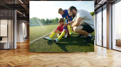 Coach providing assistance to child, boy sitting on field and feeling pain on legs, being injured during game. Concept of sport, childhood, education, achievement, active lifestyle Wall mural