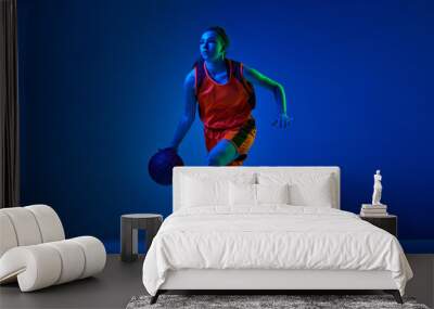 Concentrated female basketball athlete training, dribbling ball against blue studio background in neon light. Concept of professional sport, action and motion, game, competition, hobby, ad Wall mural