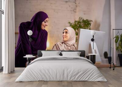 Disscussing. Happy and young two muslim women at home during lesson, studying near computer, online education. Culture, traditions, modern people. Watching at PC monitor, shopping or talking. Wall mural