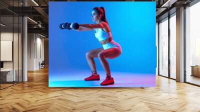 Dumbbell squats. Young sportive girl training with sports equipment isolated on gradient blue-pink studio background in neon light. Sport, action, fitness, youth concept. Wall mural