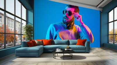 Enjoying his favorite music. Happy young stylish man in hat and sunglasses with headphones listening and smiling while standing against blue neon background Wall mural