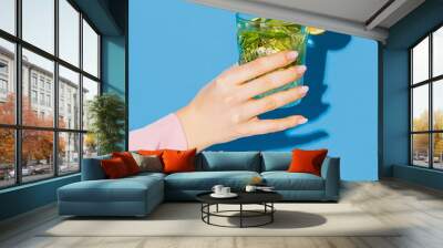 Female hand holding glass with cocktail mojito with lemon isolated on light blue neon background. Concept of taste, alcoholic drinks Wall mural