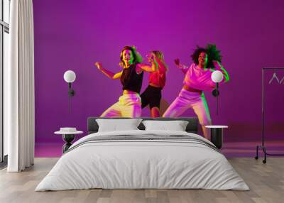 Flexible. Sportive girls dancing hip-hop in stylish clothes on purple-pink background at dance hall in green neon light. Youth culture, movement, style and fashion, action. Fashionable portrait. Wall mural