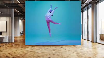 Flying high. Young and graceful ballet dancer isolated on blue studio background in neon light. Art, motion, action, flexibility, inspiration concept. Flexible caucasian ballet dancer, moves in glow. Wall mural