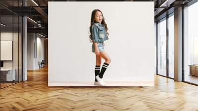 Full length portrait of cute little kid girl in stylish jeans clothes looking at camera and smiling, standing against white studio wall. Kids fashion concept Wall mural