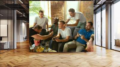 Group of excited friends playing video games at home. Caucasian male gamers or fans spending time and having fun together at home. Emotional watching gameplay. Modern technologies, friendship, weekend Wall mural