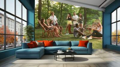 Group of happy friends eating and drinking beers at barbecue dinner on sunset time. Having meal together outdoor in a forest glade. Celebrating and relaxing. Summer lifestyle, food, friendship concept Wall mural